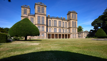 Harwick Hall lawn The image shows the lawn at Hardwick Hall, a prominent Elizabethan manor house located in Derbyshire, United Kingdom. Captured as an architectural landscape photograph, the scene is bathed in bright morning sunlight during the summer, with clear skies overhead. The historic Hardwick Hall, renowned for its symmetrical design and extensive windows, stands as a key National Trust property and well-known landmark in the region. The view highlights the manicured grounds and distinctive topiary, emphasizing the grandeur of Elizabethan architecture at this manor house in Derbyshire.